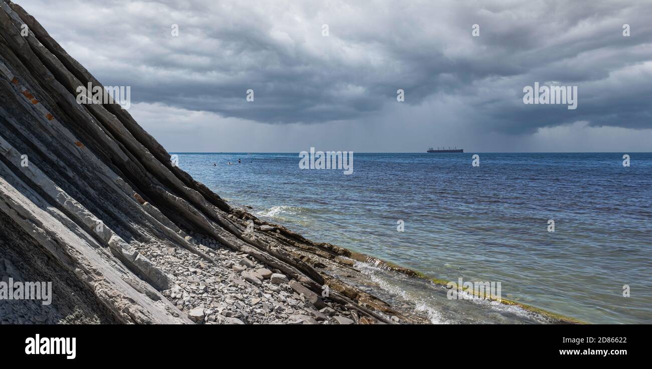 Panorama d'un paysage marin nuageux. Une plage sauvage en pierre pittoresque au pied des rochers par temps pluvieux et un bateau à l'horizon Banque D'Images