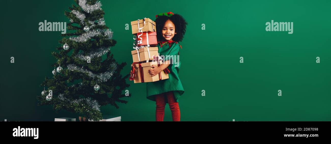 Kid holding sa gift box debout à côté d'un arbre de Noël décoré. Happy afro American girl portant un tas de boîtes cadeaux. Banque D'Images