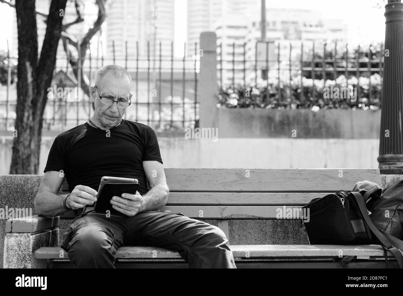 Tourisme senior Bald man using digital tablet, assis sur le banc en bois à l'encontre de la ville au parc paisible à Bangkok en Thaïlande Banque D'Images