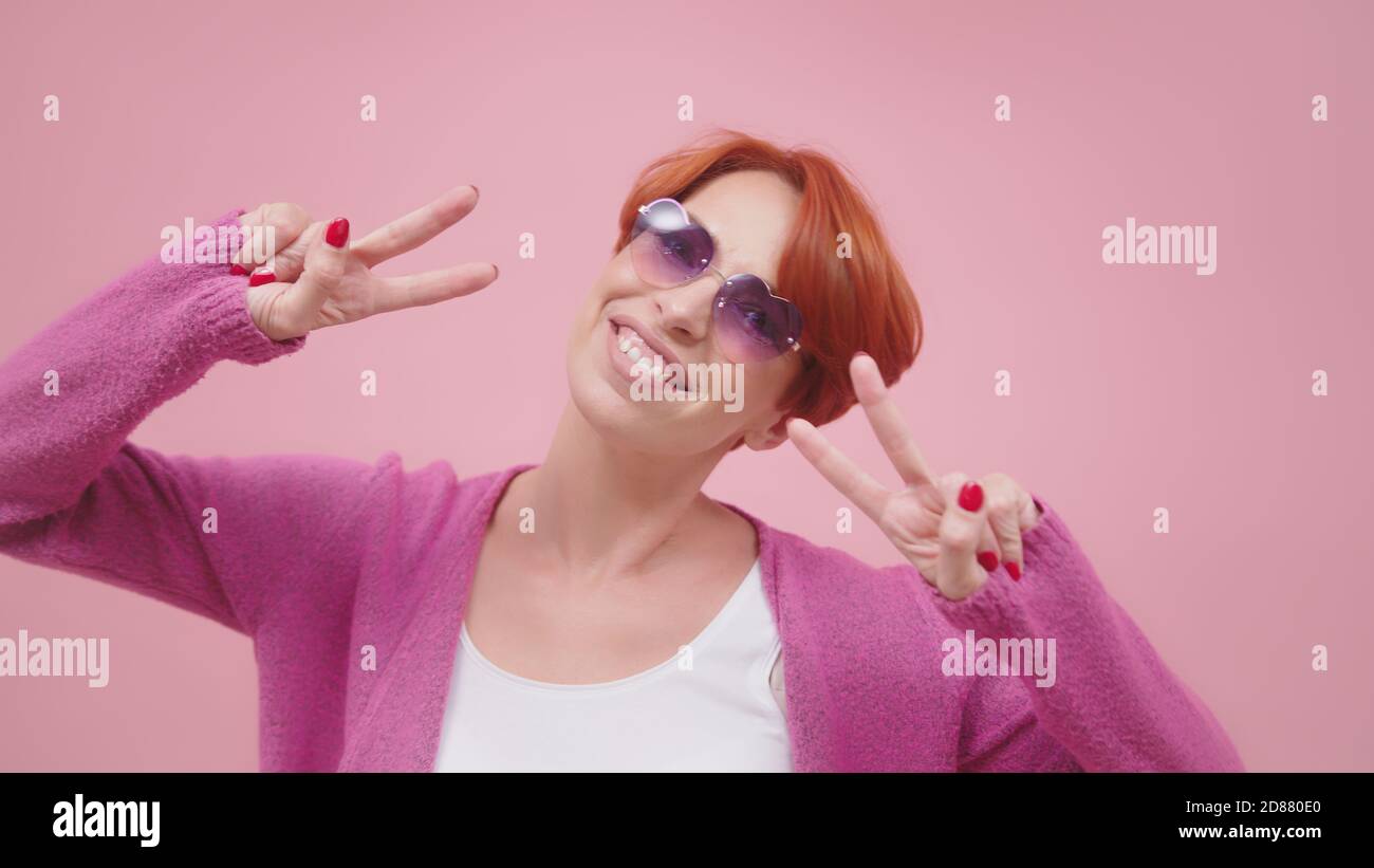 Belle femme en 40s avec des lunettes en forme de coeur rose montrant le signe de paix avec les deux mains. Isolé sur fond violet rose. Photo de haute qualité Banque D'Images