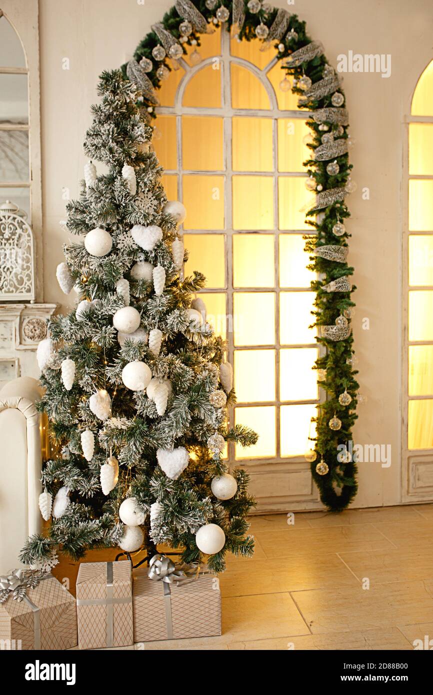 Sapin artificiel vert de Noël dans une pièce lumineuse de la maison près d'une grande fenêtre pleine hauteur avec lumière jaune. Décoré avec des boules blanches, Banque D'Images