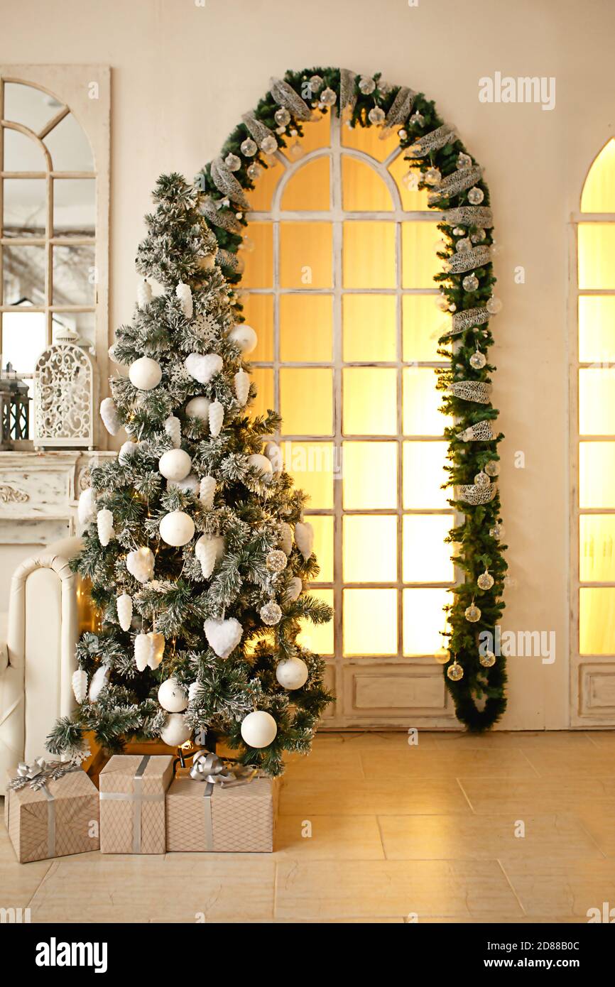 Sapin artificiel vert de Noël dans une pièce lumineuse de la maison près d'une grande fenêtre pleine hauteur avec lumière jaune. Décoré avec des boules blanches, Banque D'Images