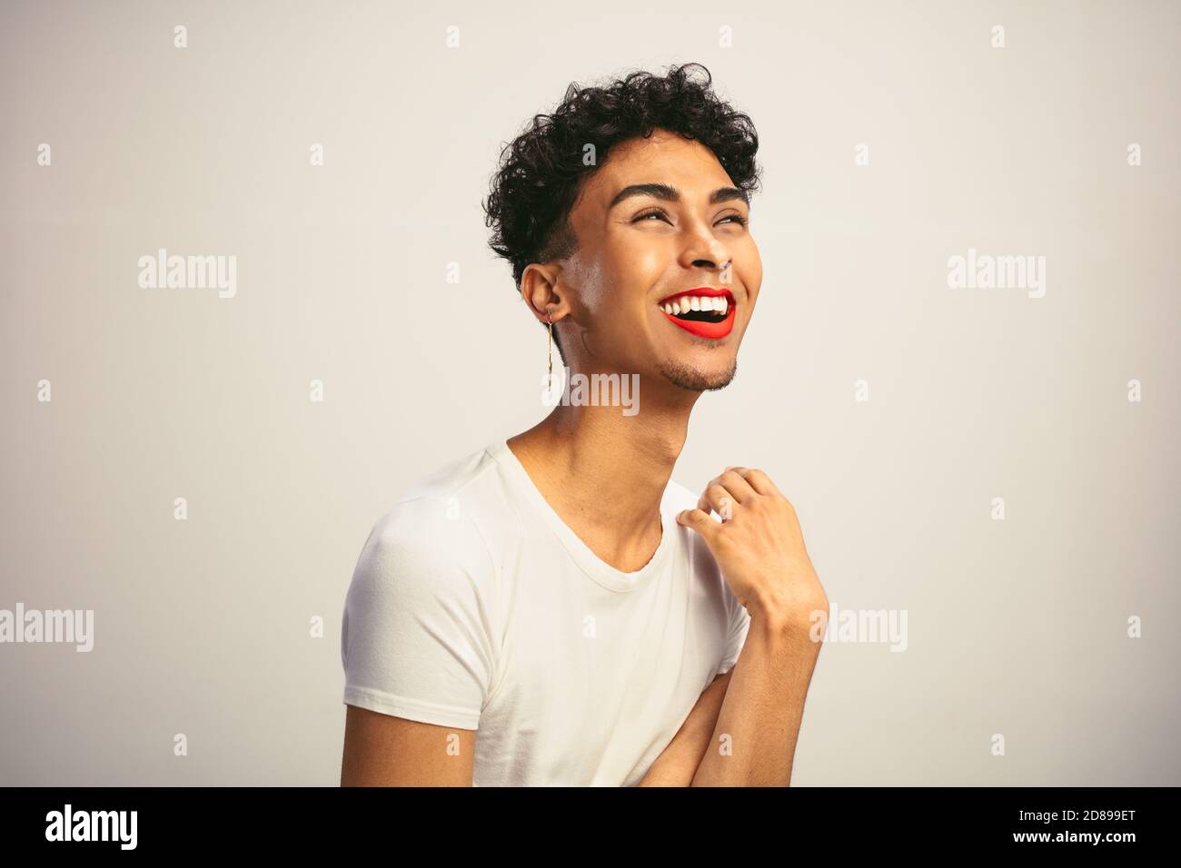 Gai gai homme avec maquillage. Homme transgenre portant un bâton rouge à lèvres et des boucles d'oreilles riant sur fond blanc. Banque D'Images