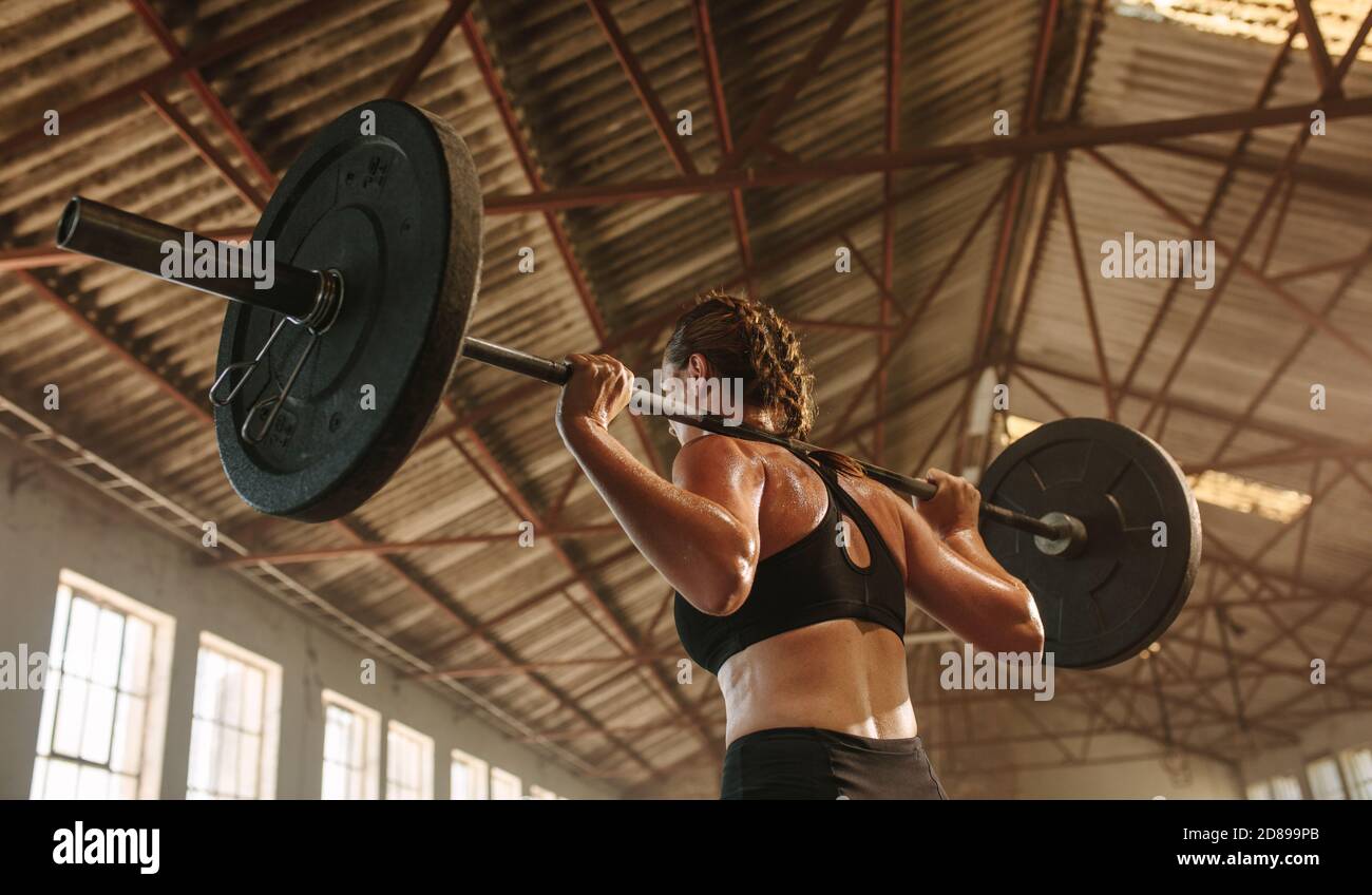 Femme forte s'exerçant avec barbell. FIT femme s'entraîner avec des poids lourds à la salle d'entraînement croisée à l'ombre de l'usine. Banque D'Images
