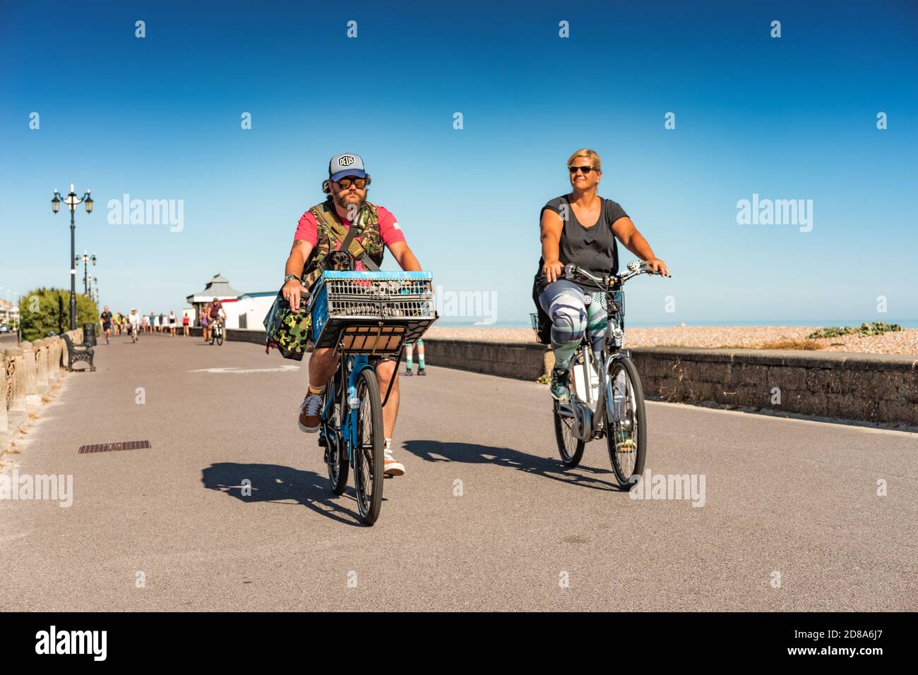 Un jeune couple adulte qui fait du vélo le long de la promenade de Worthing avec un petit chien dans un panier sous le ciel bleu les jours d'été. Banque D'Images