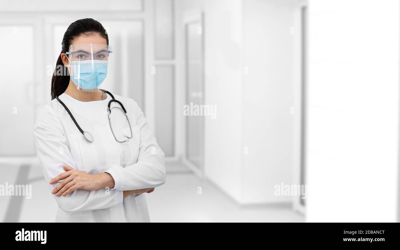 Portrait de face d'une femme médecin de race mixte portant un écran de protection et un masque chirurgical, debout avec les bras repliés à l'hôpital de couloir Banque D'Images