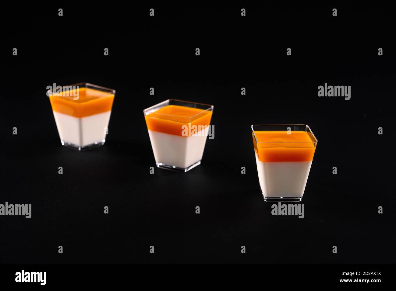 Dessert sucré décoré avec une garniture orange vif, isolé sur fond de studio noir. Vue de face de trois petits verres carrés avec une délicieuse panna cotta laiteuse. Concept alimentaire. Banque D'Images