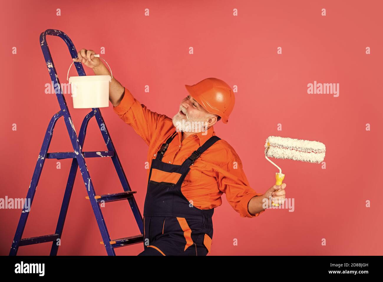 travail en atelier de réparation. peintre homme senior utilise le rouleau sur l'échelle. peinture du mur en rose. peintre professionnel en vêtements de travail. mur de peinture ouvrier dans la salle. peinture décorateur mâle avec rouleau. Banque D'Images