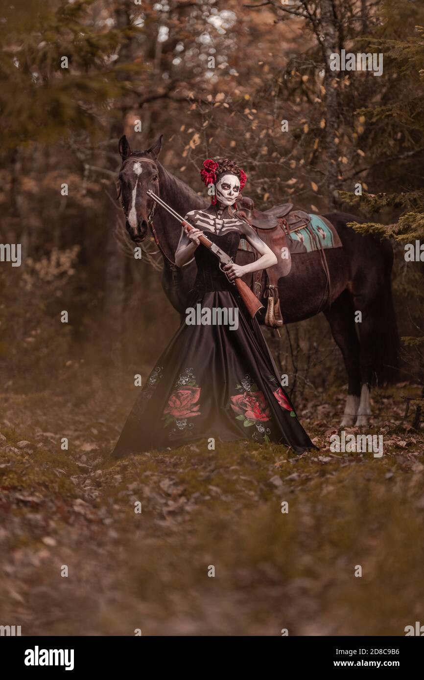 Jeune femme habillée comme le symbole mexicain de l'époque de la calavera morte dans une robe noire posant dans la forêt avec un cheval Banque D'Images