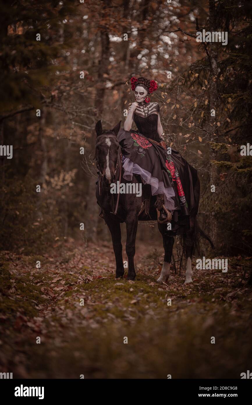 Jeune femme habillée comme le symbole mexicain de l'époque de la calavera morte dans une robe noire posant dans la forêt avec un cheval Banque D'Images