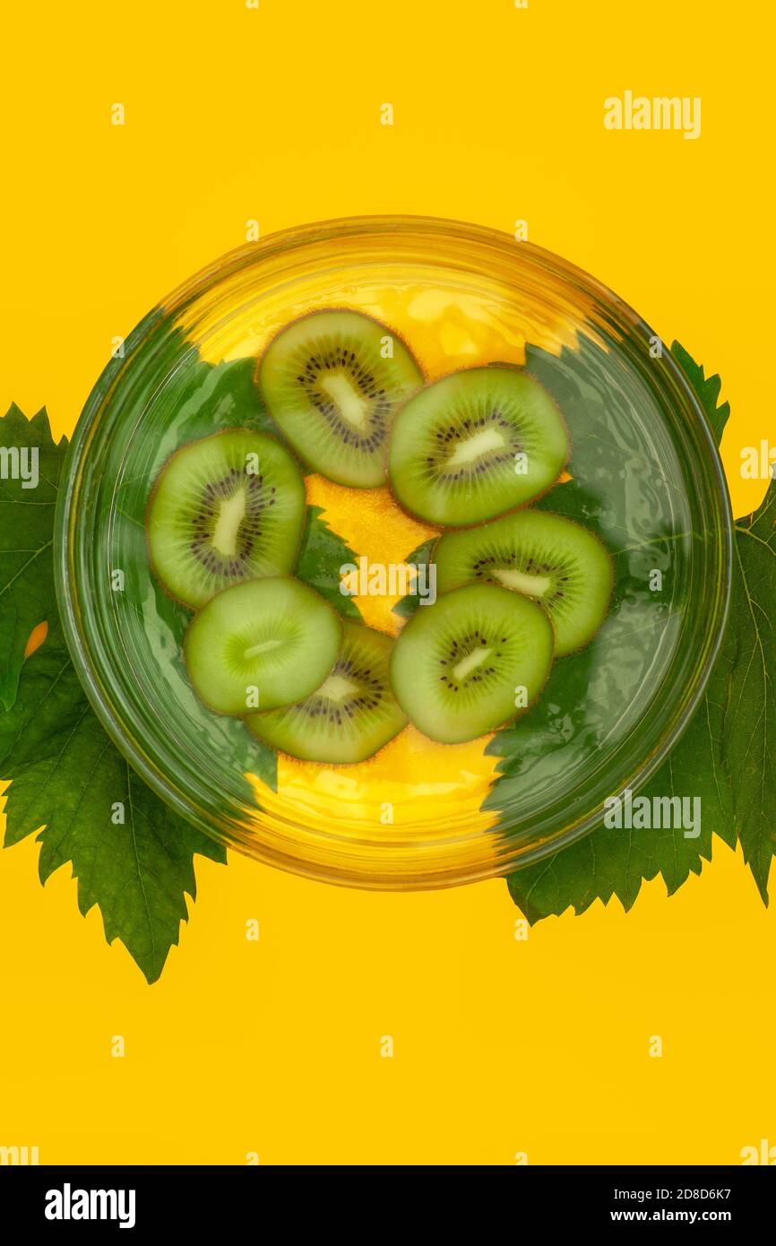 Kiwi dans un bol en verre sur fond jaune décoré avec feuilles de raisin vert Banque D'Images