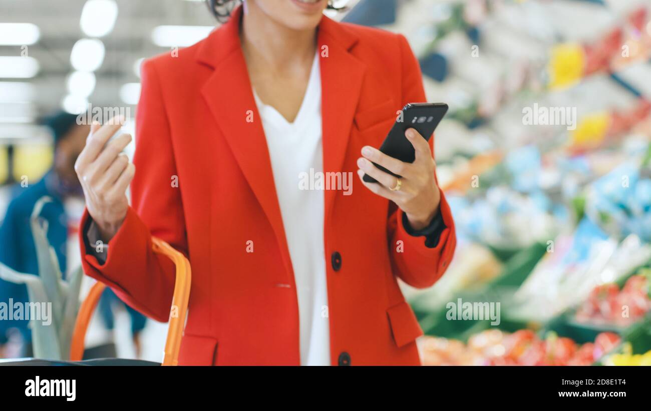 Au supermarché: Belle jeune femme utilise un smartphone tout en se tenant à la section des produits frais du magasin. En arrière-plan autres clients Banque D'Images