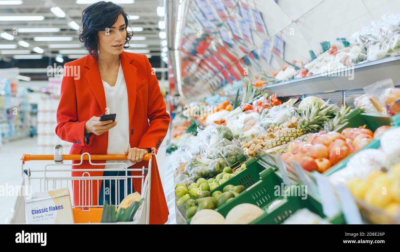 Au supermarché: Belle jeune femme marche à travers la section des produits frais, choisit des légumes et pousser le panier. Le client utilise un smartphone Banque D'Images