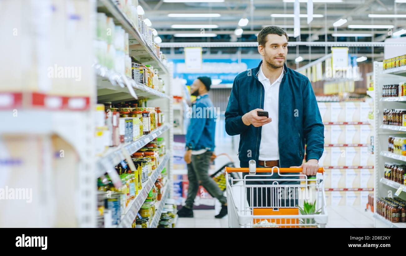 Au supermarché : Handsome Man utilise un smartphone et navigue à travers la tablette de marchandises en conserve. Il est debout avec le chariot dans la section des marchandises en conserve. Banque D'Images