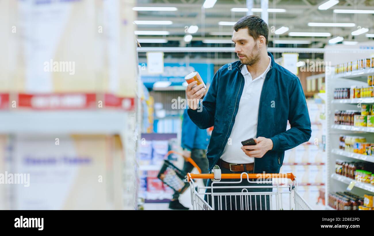 Au supermarché : Handsome Man utilise un smartphone et navigue à travers la tablette de marchandises en conserve. Il est debout avec le chariot dans la section des marchandises en conserve. Banque D'Images