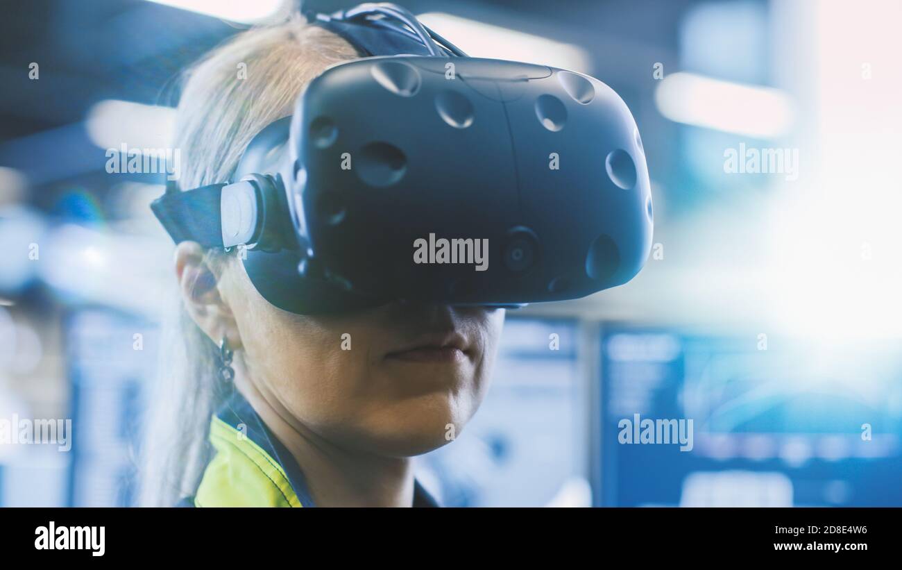 À l'usine : une femme ingénieure portant un casque de réalité virtuelle travaille sur un projet, déplaçant sa tête. Dans les écrans arrière-plan et industriel Banque D'Images