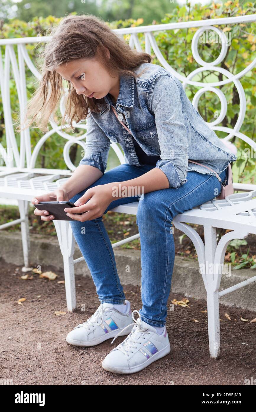 Belle jeune fille d'âge en utilisant un smartphone tout en étant assis sur le banc dans le parc, jeans vêtements modernes Banque D'Images