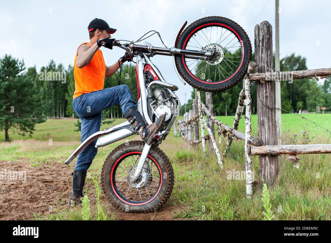 Homme pilote avec entraînement de motocycle d'essai dans la zone de campagne, levant le vélo sur la roue arrière, penché contre la clôture en bois Banque D'Images