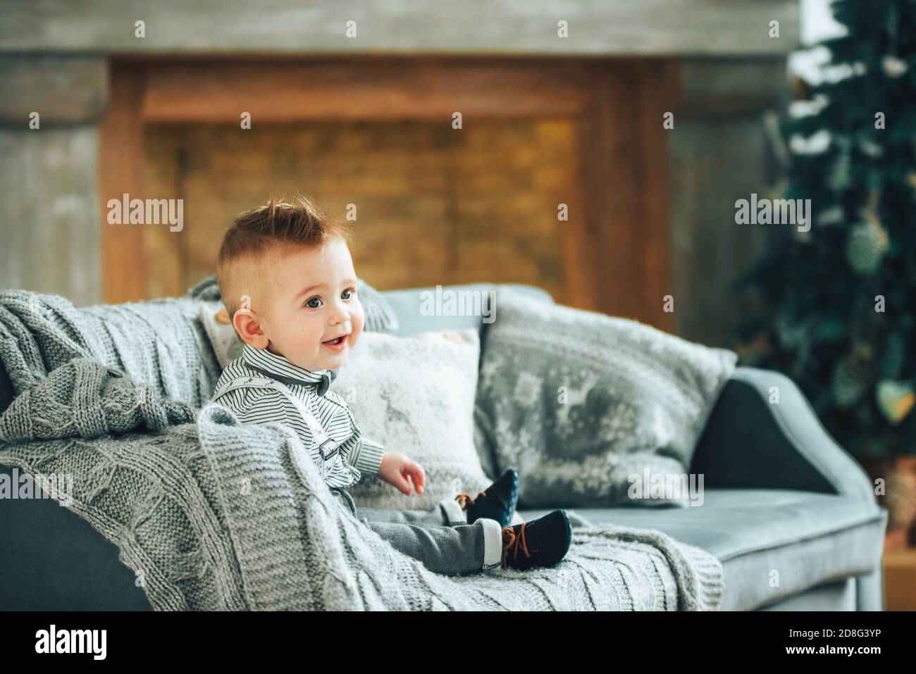 Mignon garçon d'un an assis sur le canapé recouvert d'une couverture grise dans la chambre avec décoration de Noël. Un petit homme souriant. Portrait des enfants. Placer pour le texte Banque D'Images