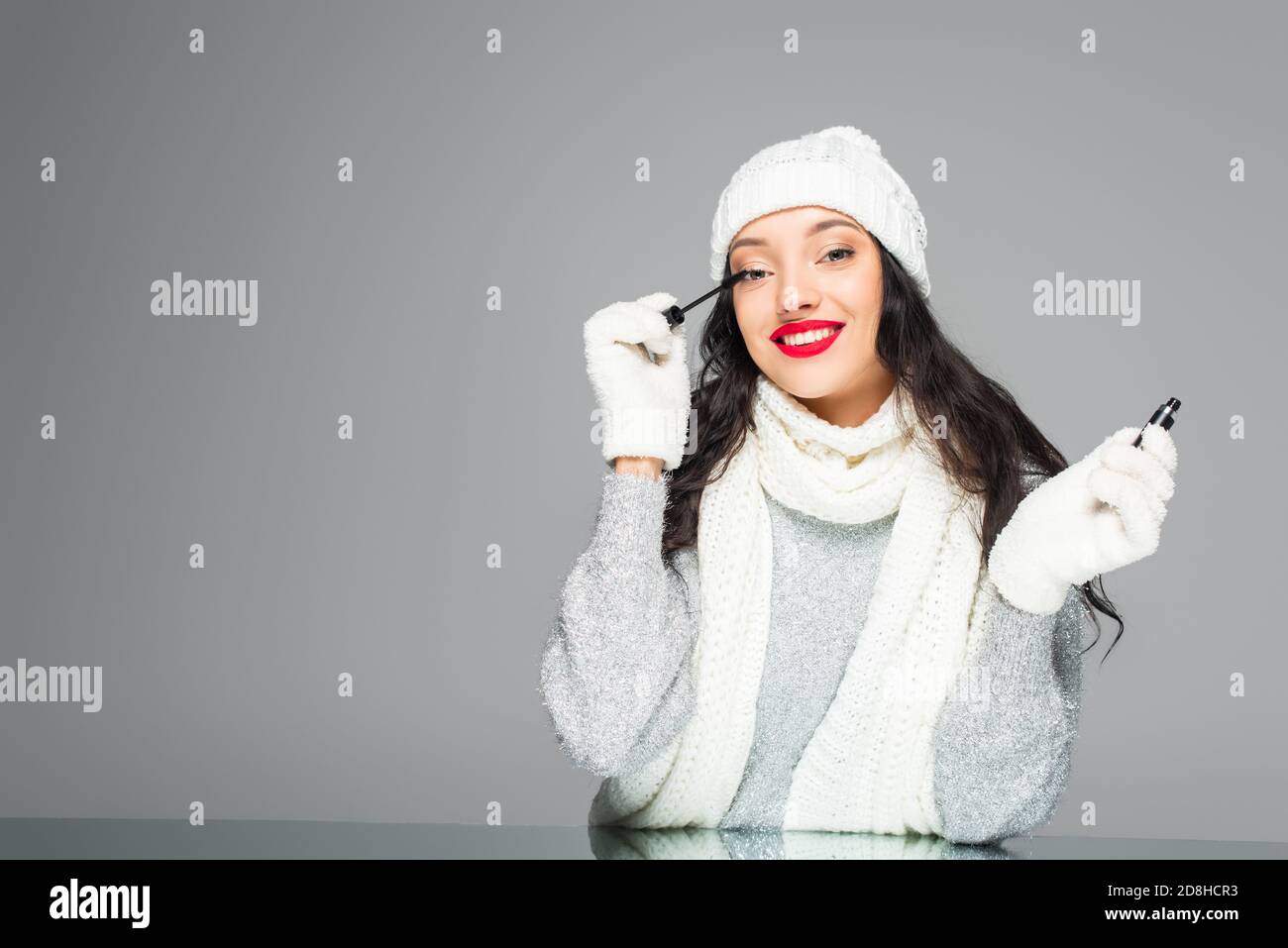 femme heureuse et brune en tenue d'hiver appliquant la mascara isolée en gris Banque D'Images
