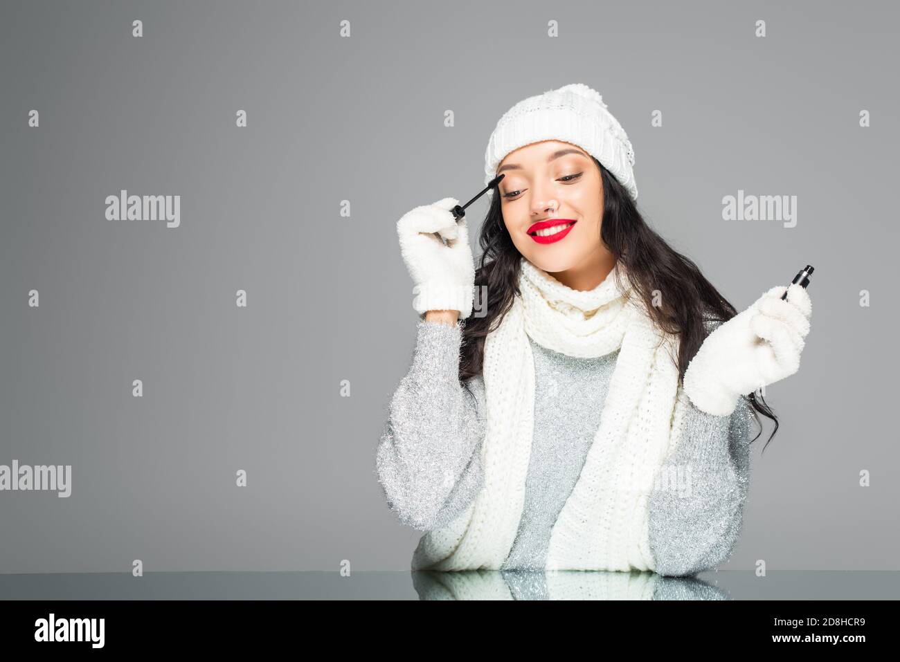 femme joyeuse et brune en tenue d'hiver appliquant la mascara isolée en gris Banque D'Images