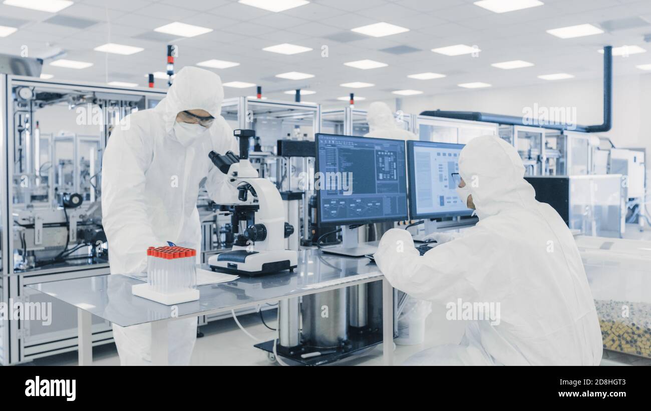 Équipe de chercheurs scientifiques en costumes stériles travaillant avec des ordinateurs, sous microscope et machines industrielles modernes dans le laboratoire Banque D'Images