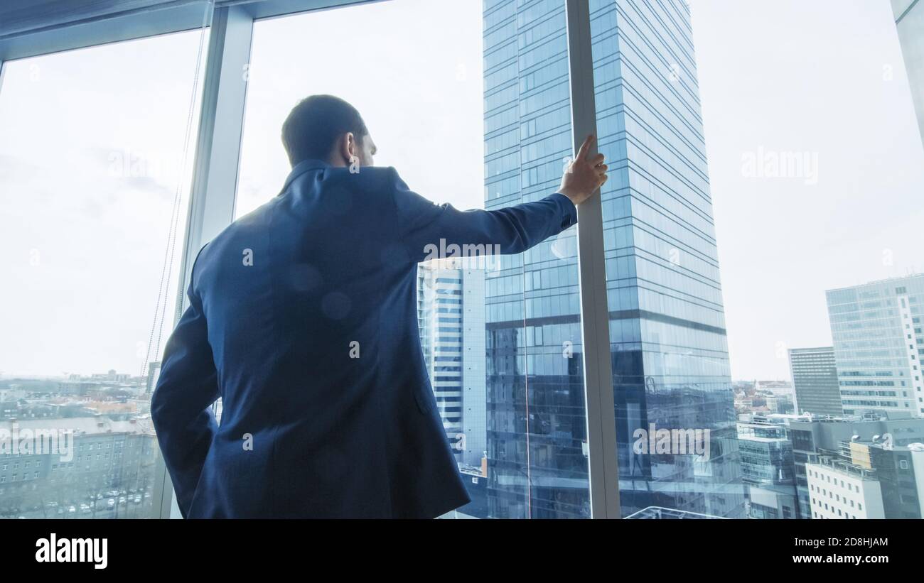 Homme d'affaires sérieux, vêtu d'un costume debout dans son bureau, regardant par la fenêtre et envisageant le prochain contrat Big Business. Ville principale Banque D'Images