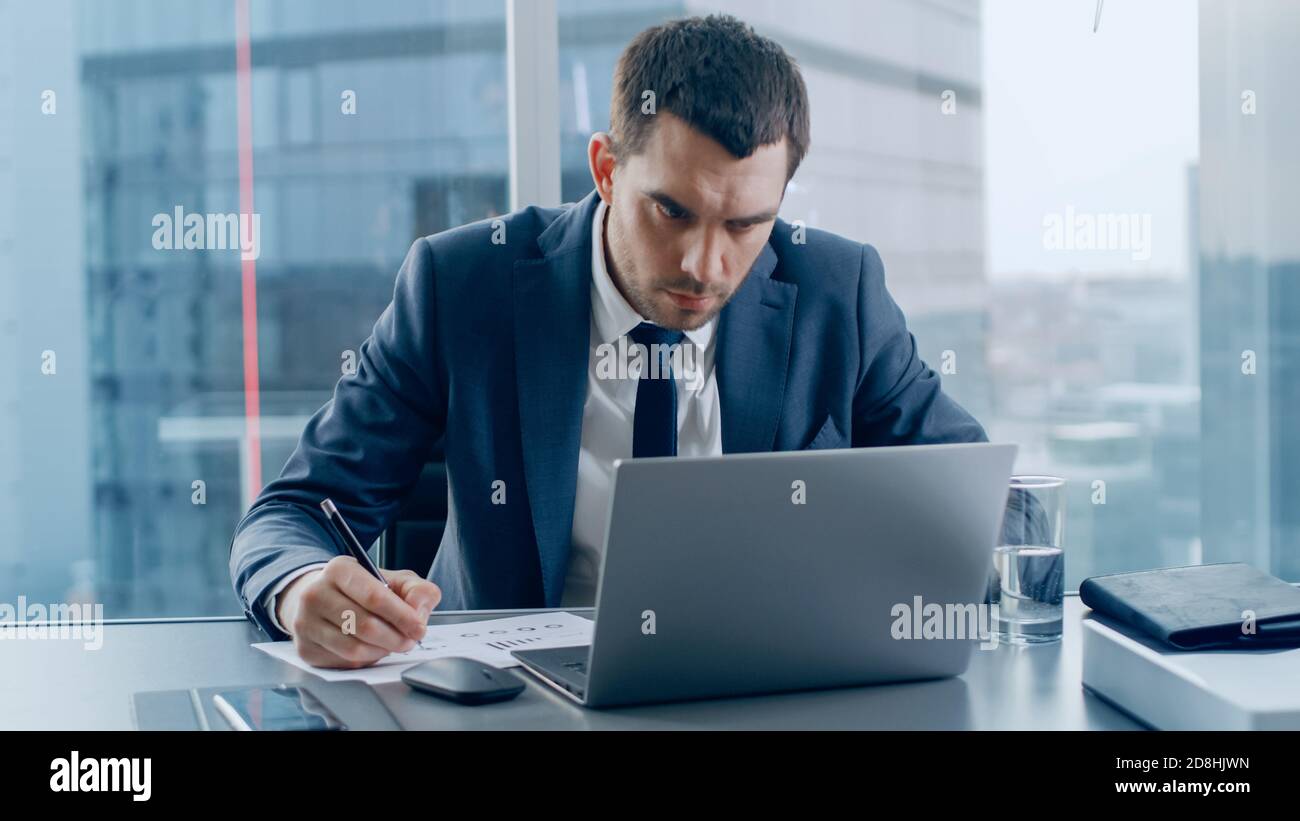 Homme d'affaires prospère assis à son bureau, travaillant avec des papiers, signant violemment des documents et regardant l'ordinateur portable. Dans la fenêtre Big City Business Banque D'Images