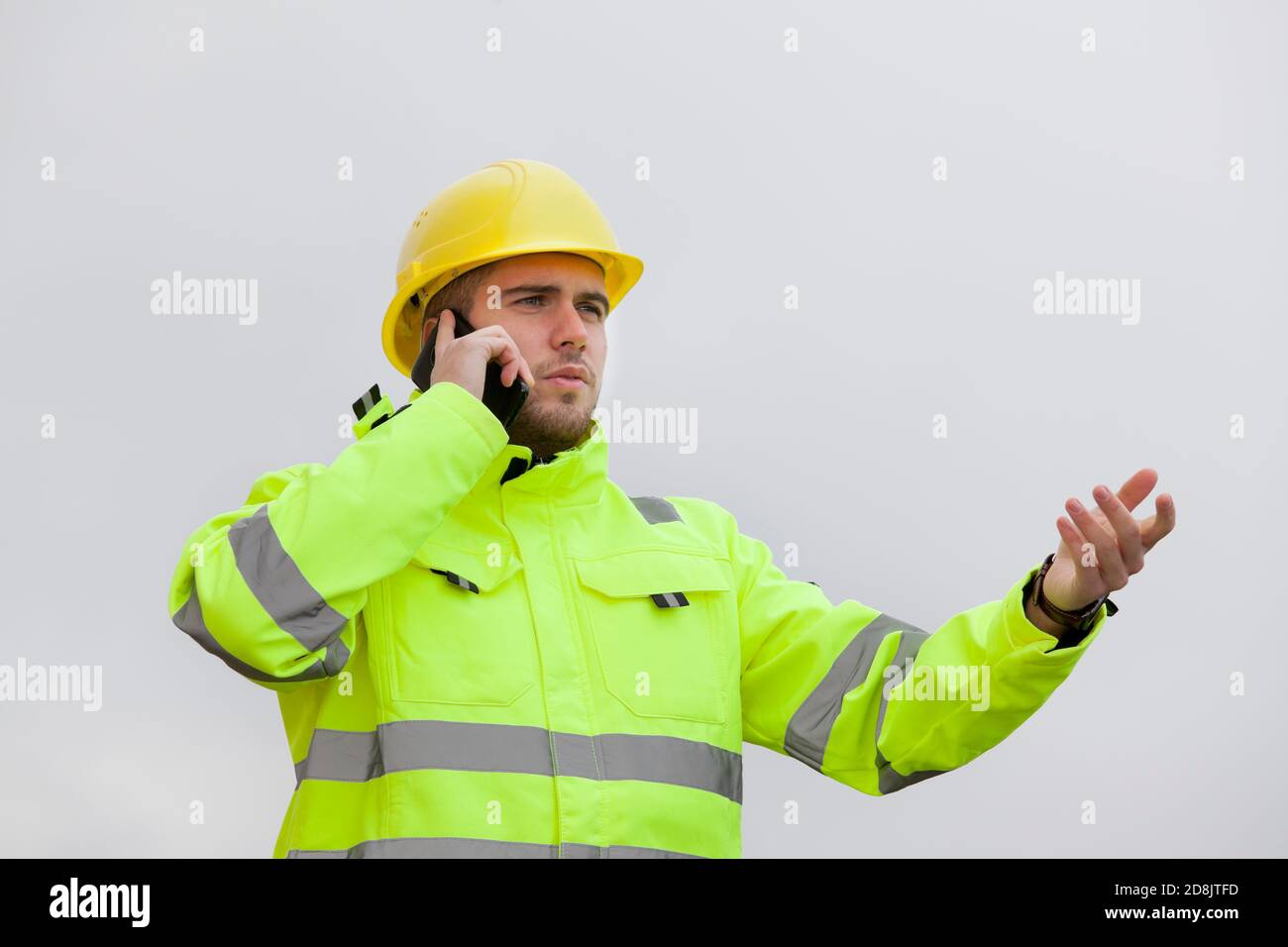 Jeune ingénieur ou travailleur avec des vêtements de travail de protection verte parler sur un téléphone portable devant un ciel lumineux Banque D'Images