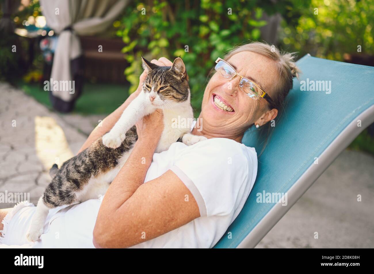 Heureuse femme âgée souriante dans des verres se détendre dans le jardin d'été à l'extérieur embrassant domestique tabby chat. Concept de vieux retraités et animaux de compagnie Banque D'Images