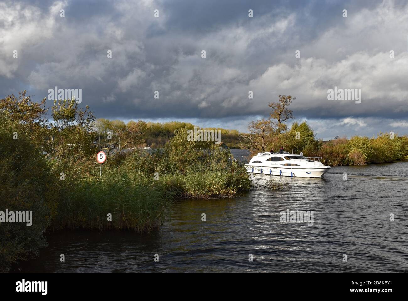 Un petit bateau émerge de Wroxham Broad pour rejoindre la rivière Bure alors que les nuages de tempête se rassemblent au-dessus. Un panneau de 5 mille par heure est vu dans les buissons, en Angleterre. Banque D'Images