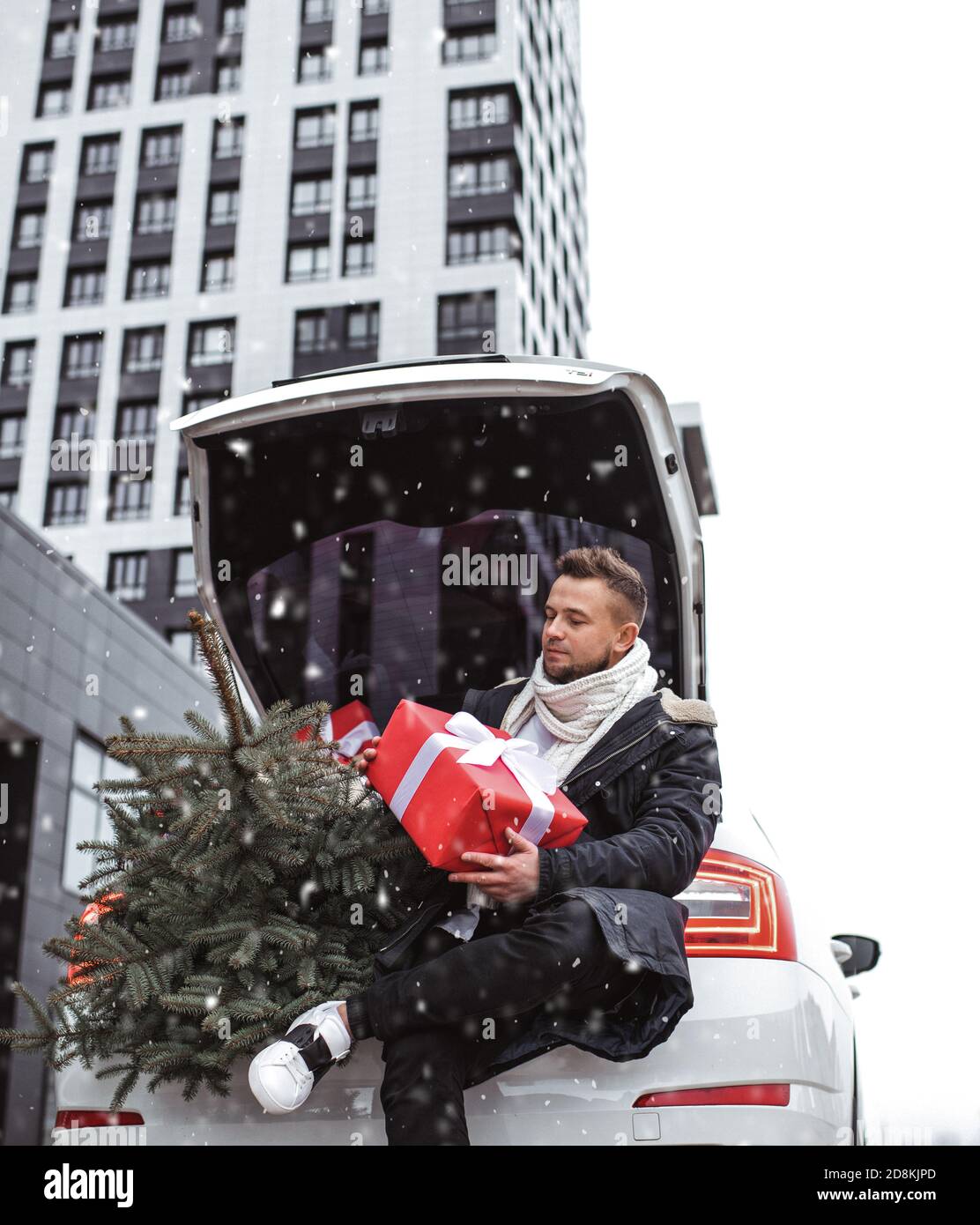 Beau homme en écharpe tricotée et gants noirs tenant une boîte cadeau, attend son bien-aimé. Fond urbain. Attente près de la voiture en chute de neige. Banque D'Images