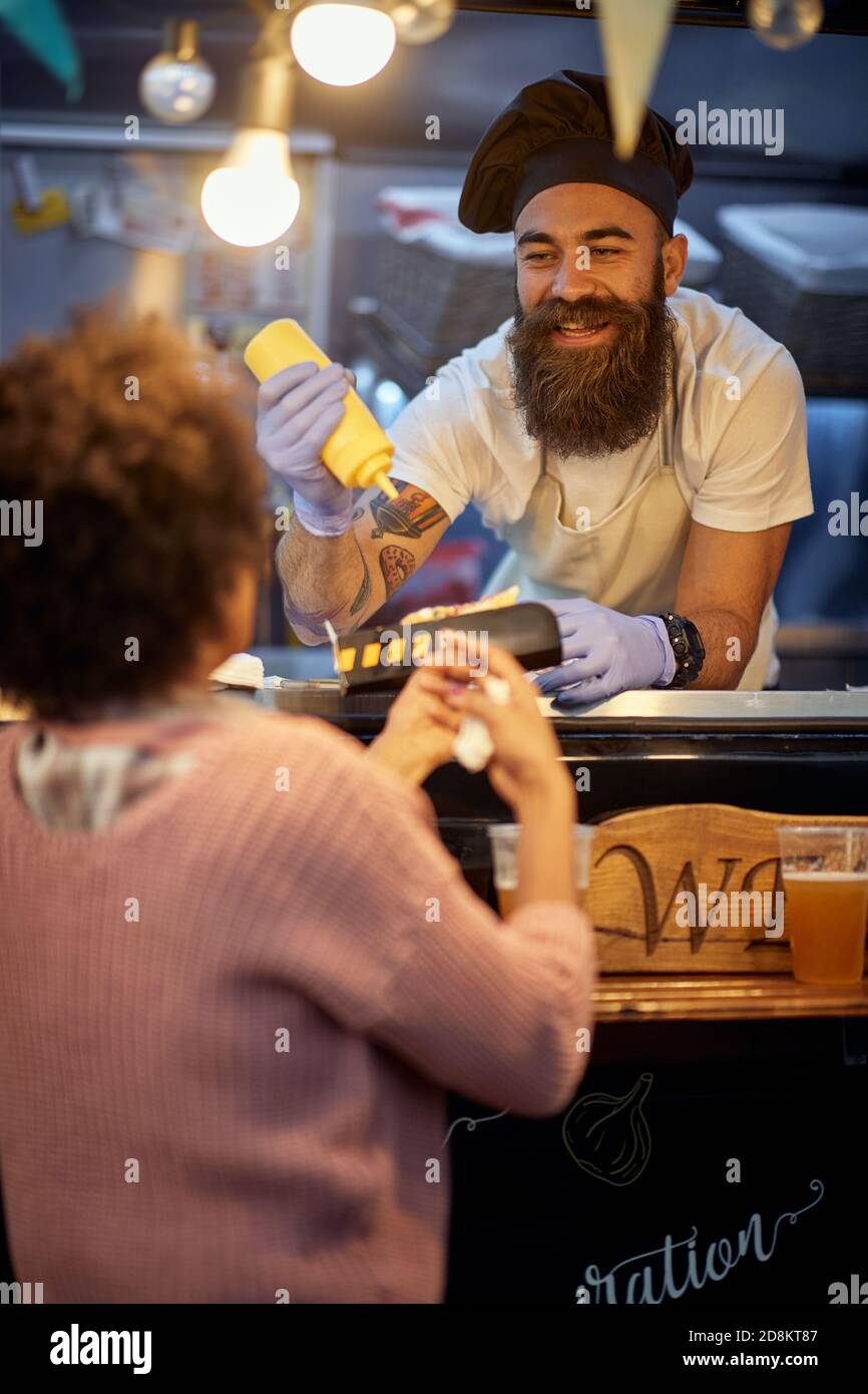 employé de sexe masculin dans le service de restauration rapide souriant et parlant à un client tout en ajoutant de la moutarde dans un sandwich Banque D'Images