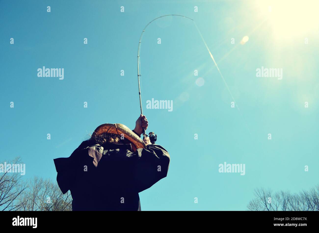 Homme portant un chapeau de cow-boy, dos tourné vers l'appareil photo. Tenant un poteau de pêche courbé en haut, avec la ligne attrapant l'éblouissement du soleil. Banque D'Images