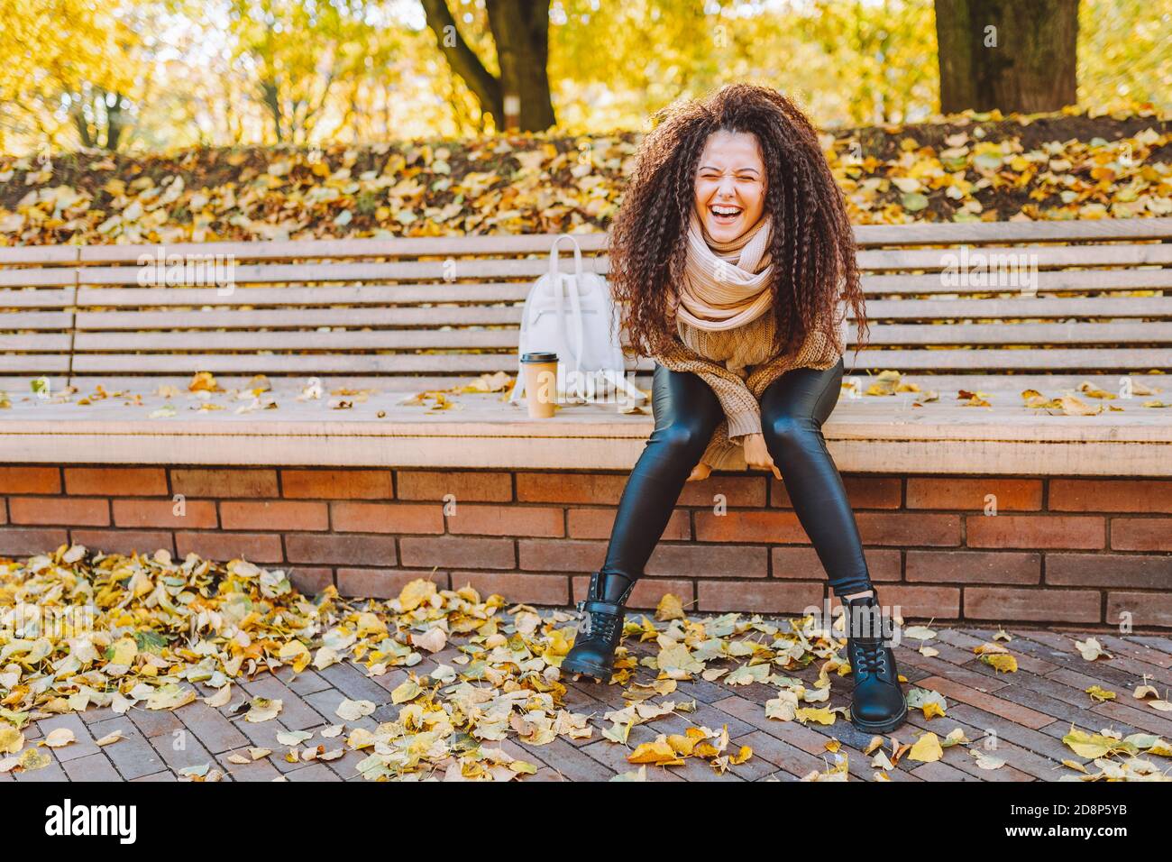 Positive afro cheveux femme avec beau sourire portant pull tricoté et le foulard s'assoient sur le banc dans le parc d'automne sous le soleil journée avec café Banque D'Images