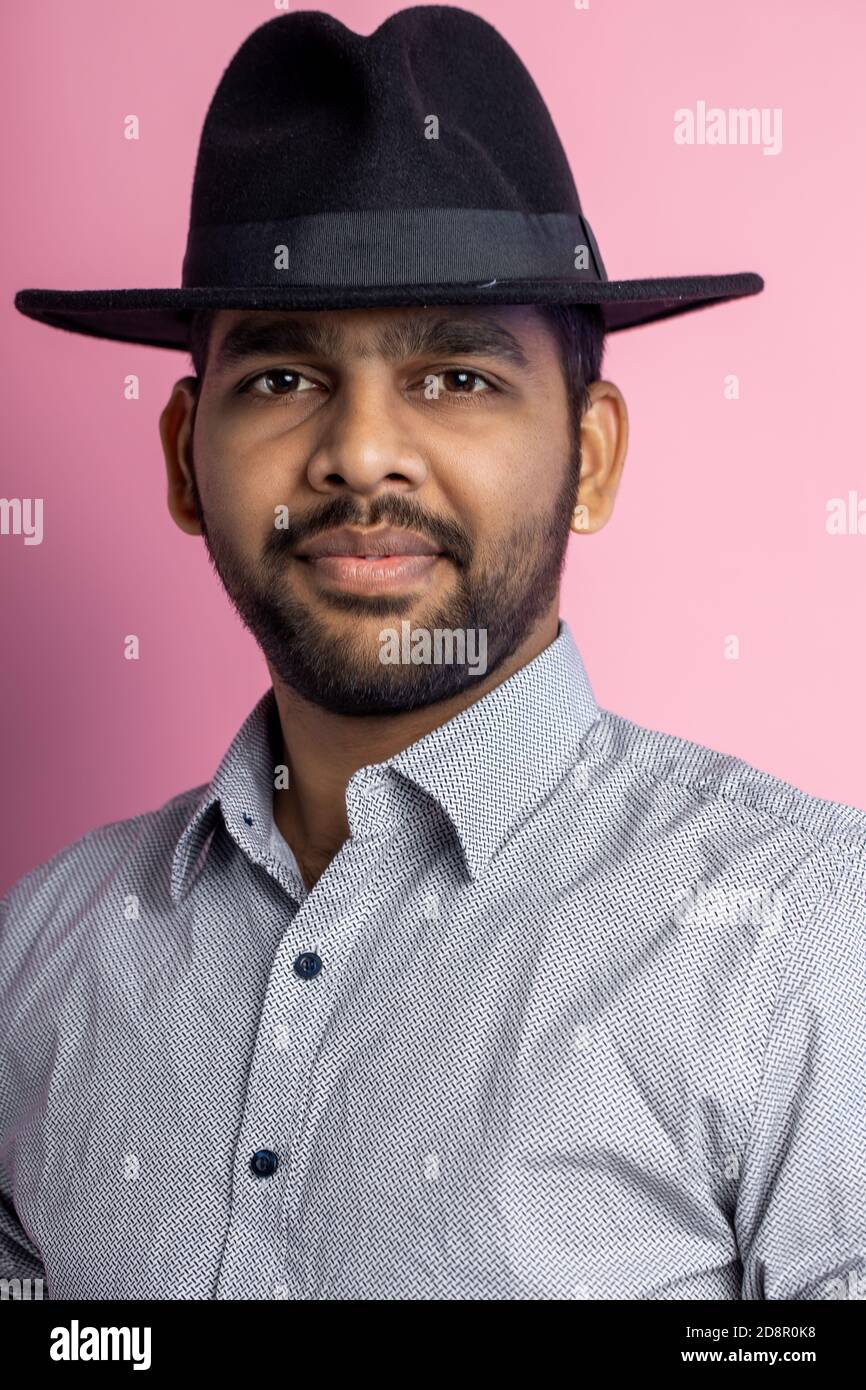 Gros plan de jeune homme indien joyeux et heureux avec barbe de 3 jours, portant une chemise décontractée, un chapeau noir, regardant avec le sourire et pointant sur le côté avec l'index Banque D'Images