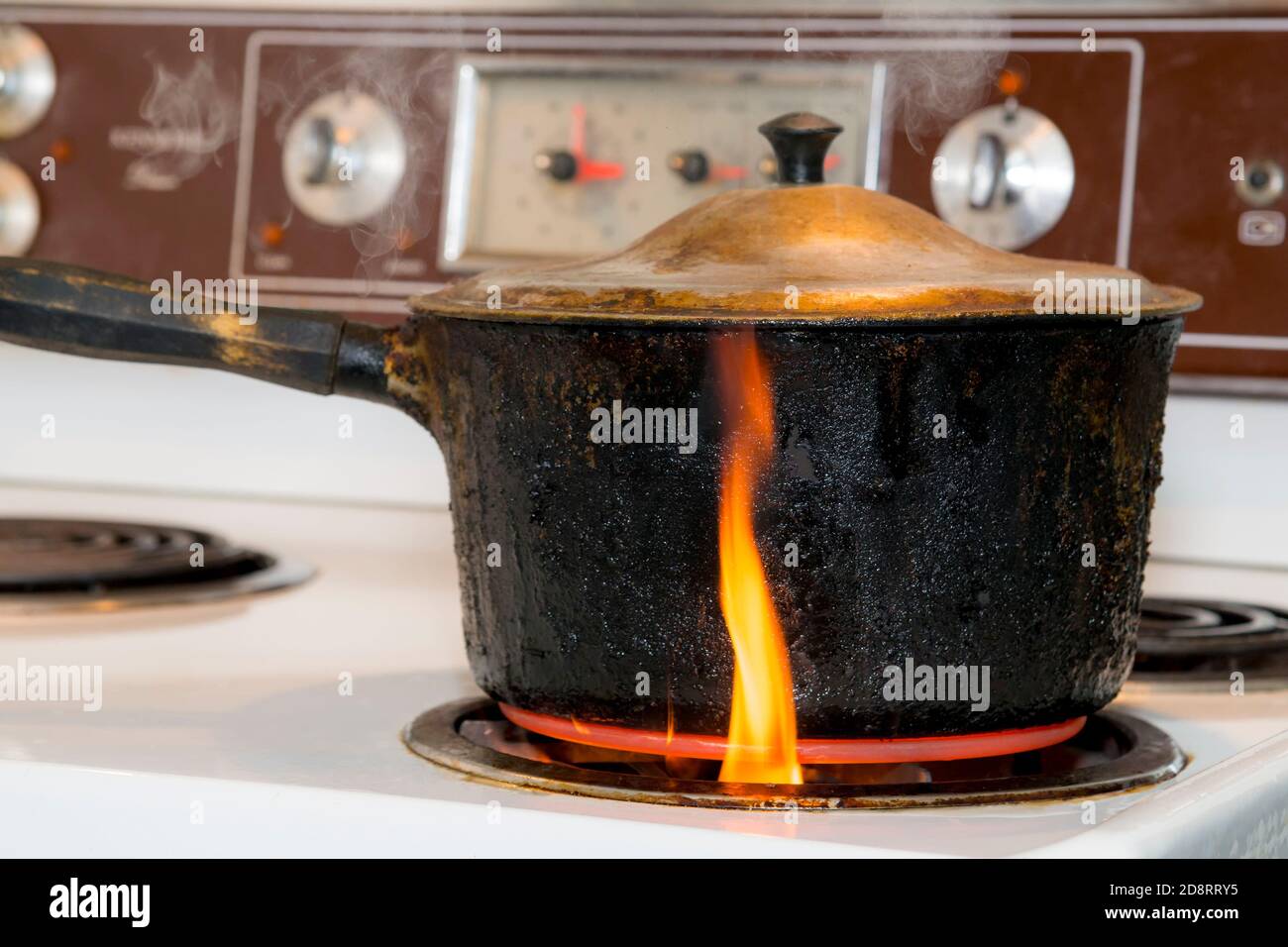 Une vieille casserole couverte, charrée, noircie brûlante sur un brûleur sur une cuisinière électrique. La petite flamme se trouve à l'extérieur du pot sortant du brûleur. Banque D'Images