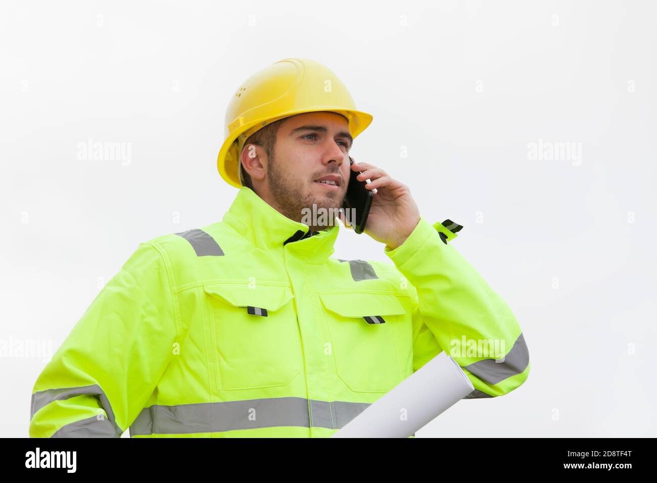 Jeune ingénieur ou travailleur avec des vêtements de travail de protection verte parler sur un téléphone portable devant un ciel lumineux Banque D'Images