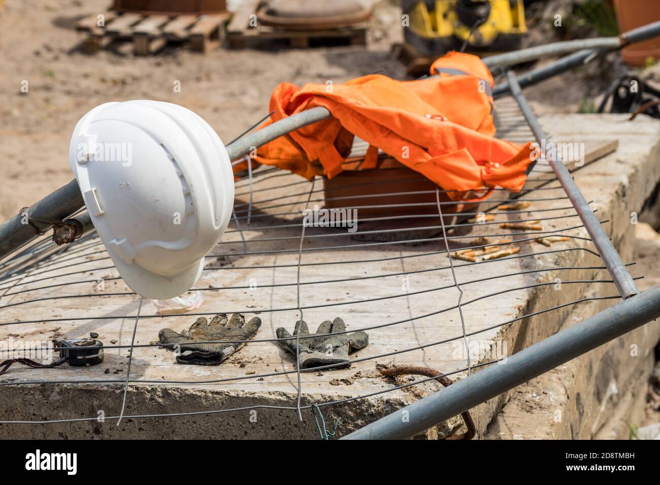 casque de construction et gilet réfléchissant couchés sur une maille métallique et une dalle de béton, en arrière-plan un chantier de construction Banque D'Images