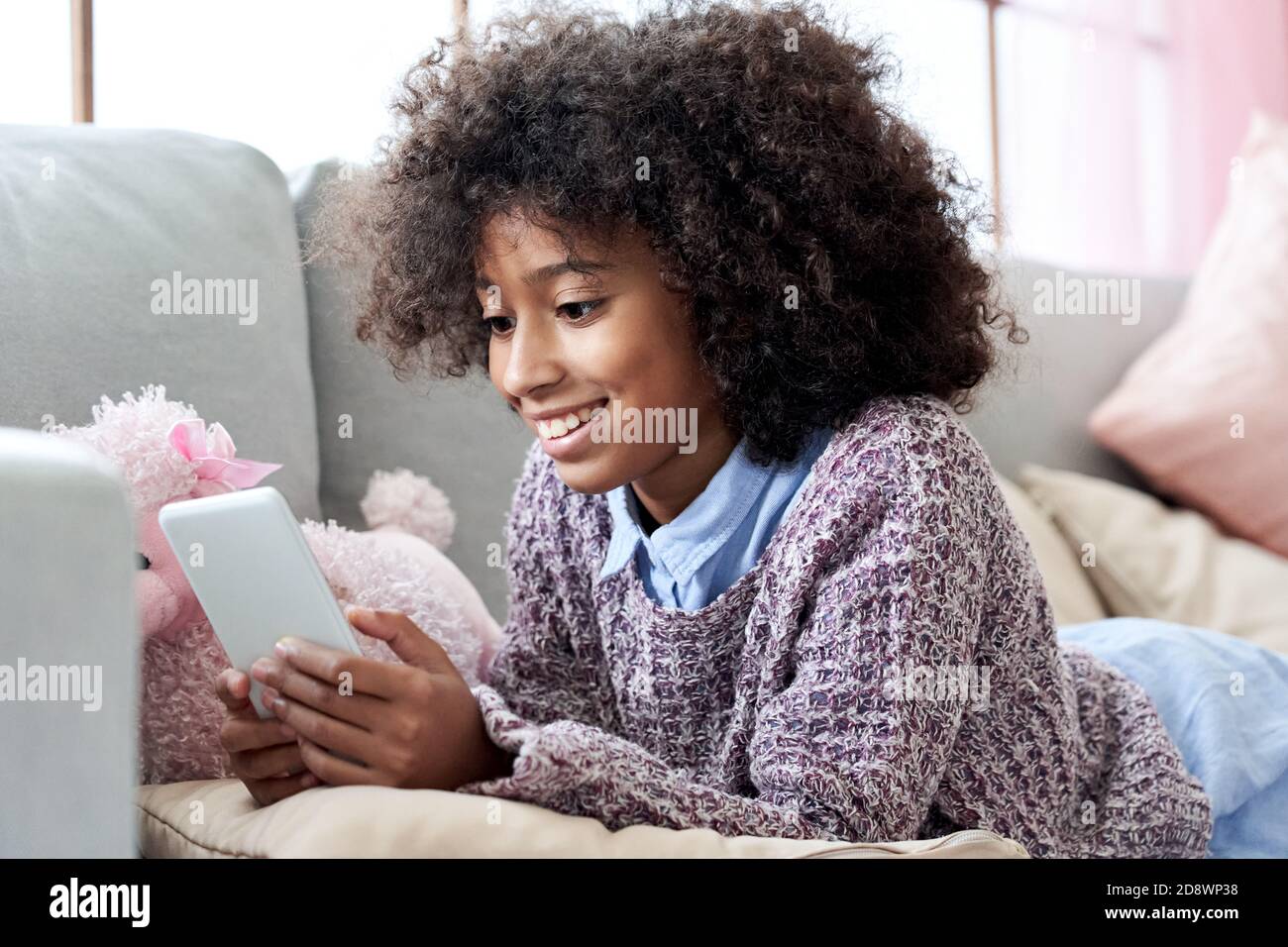 Une fillette africaine heureuse utilisant des applications mobiles sur le téléphone se détendant sur le canapé à la maison. Banque D'Images