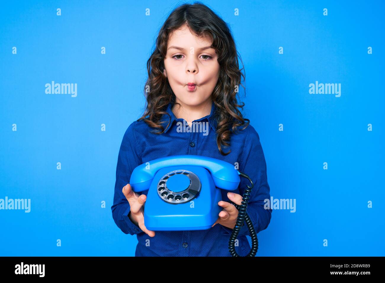 Mignon petit garçon hispanique avec de longs cheveux tenant téléphone vintage faisant le visage de poisson avec la bouche et les yeux de accroupi, fou et comique. Banque D'Images