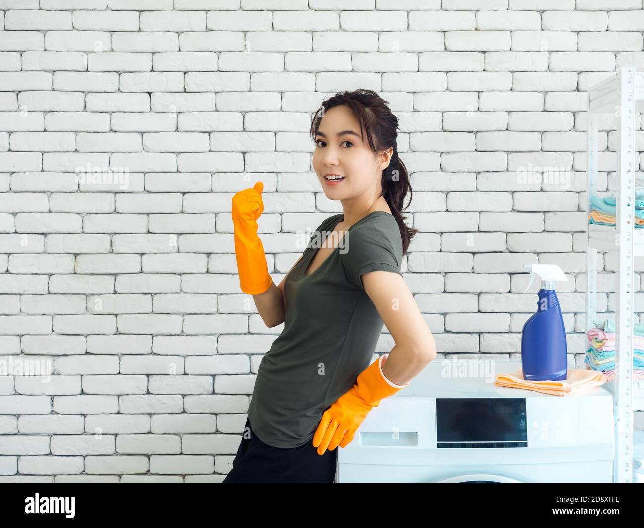 Bonne femme asiatique souriante, femme au foyer portant des gants en caoutchouc orange debout et levant la main dans le geste gagnant et célébrant le succès près de laver ma Banque D'Images