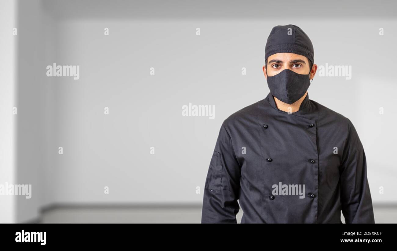 Chef à l'époque des coronavirus. Homme en uniforme de cuisinier avec masque de protection du visage en tissu noir, fond intérieur de la pièce. COVID19 sécurité dans cuisine restaurant f Banque D'Images