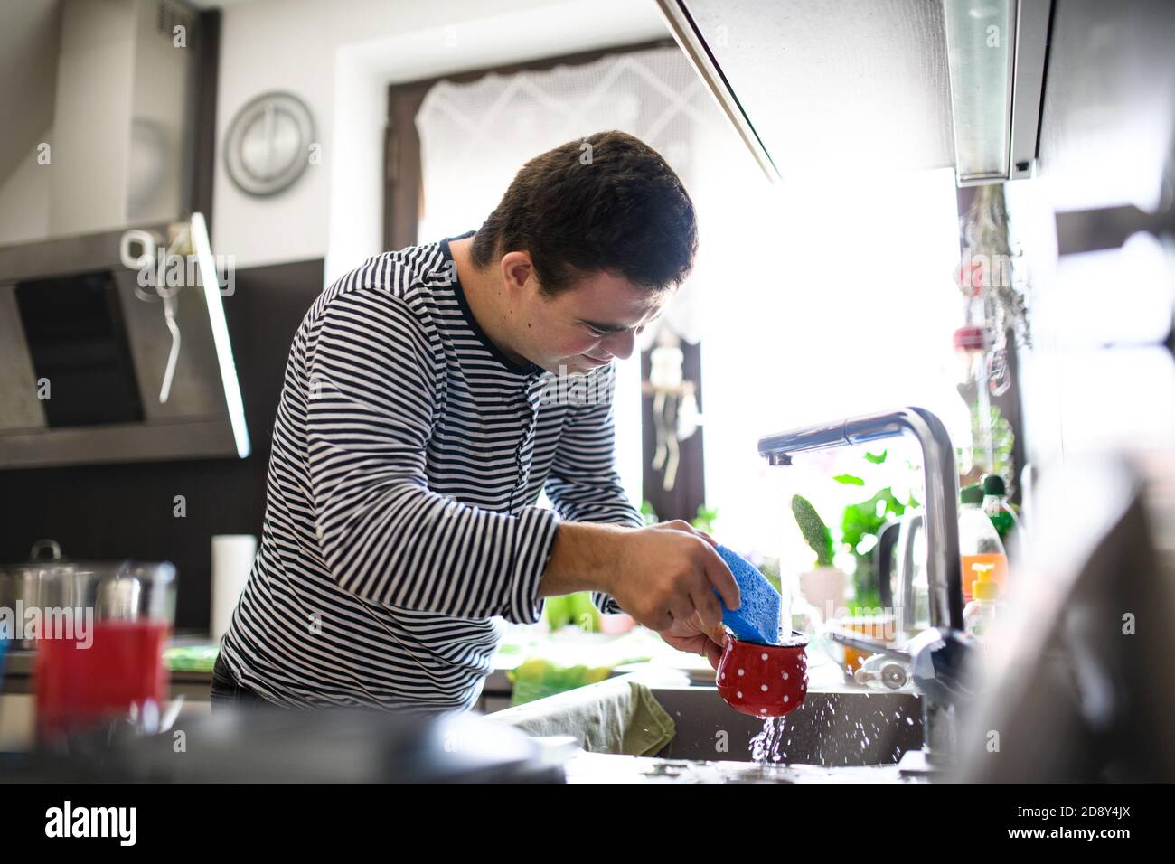Syndrome de Down homme adulte debout à l'intérieur dans la cuisine à la maison, laver la vaisselle. Banque D'Images