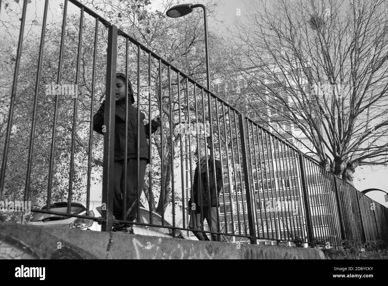 Père marchant rue avec enfant regardant à travers les rails de fer dans le cadre urbain avec bloc d'appartement en arrière-plan, B/W, Hackney, Londres, Angleterre, Royaume-Uni Banque D'Images