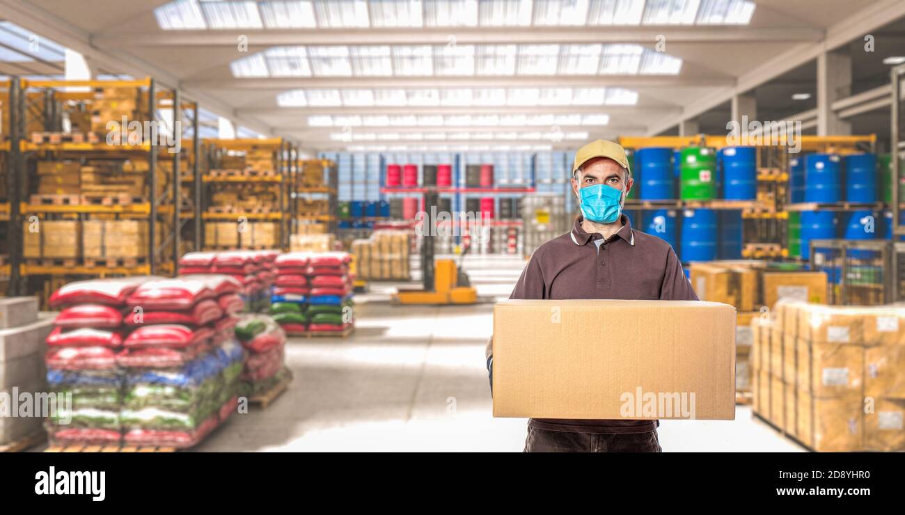 homme avec masque et colis, intérieur d'un entrepôt industriel où différentes marchandises sont stockées. Banque D'Images
