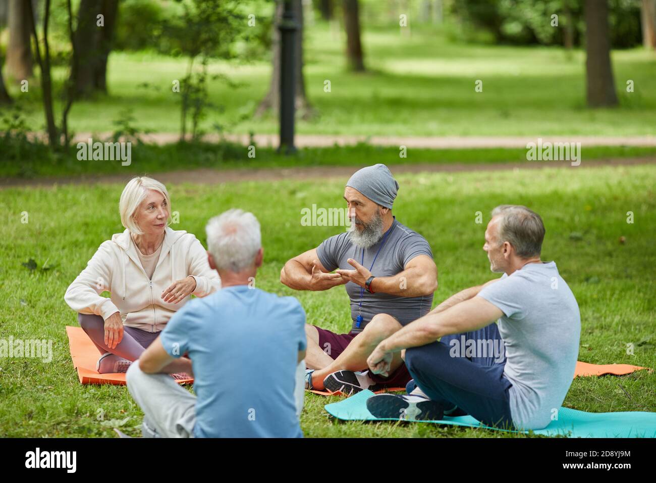 Groupe de personnes âgées actives rassemblées dans le parc de la ville assis sur des tapis à l'écoute de leur entraîneur de yoga Banque D'Images