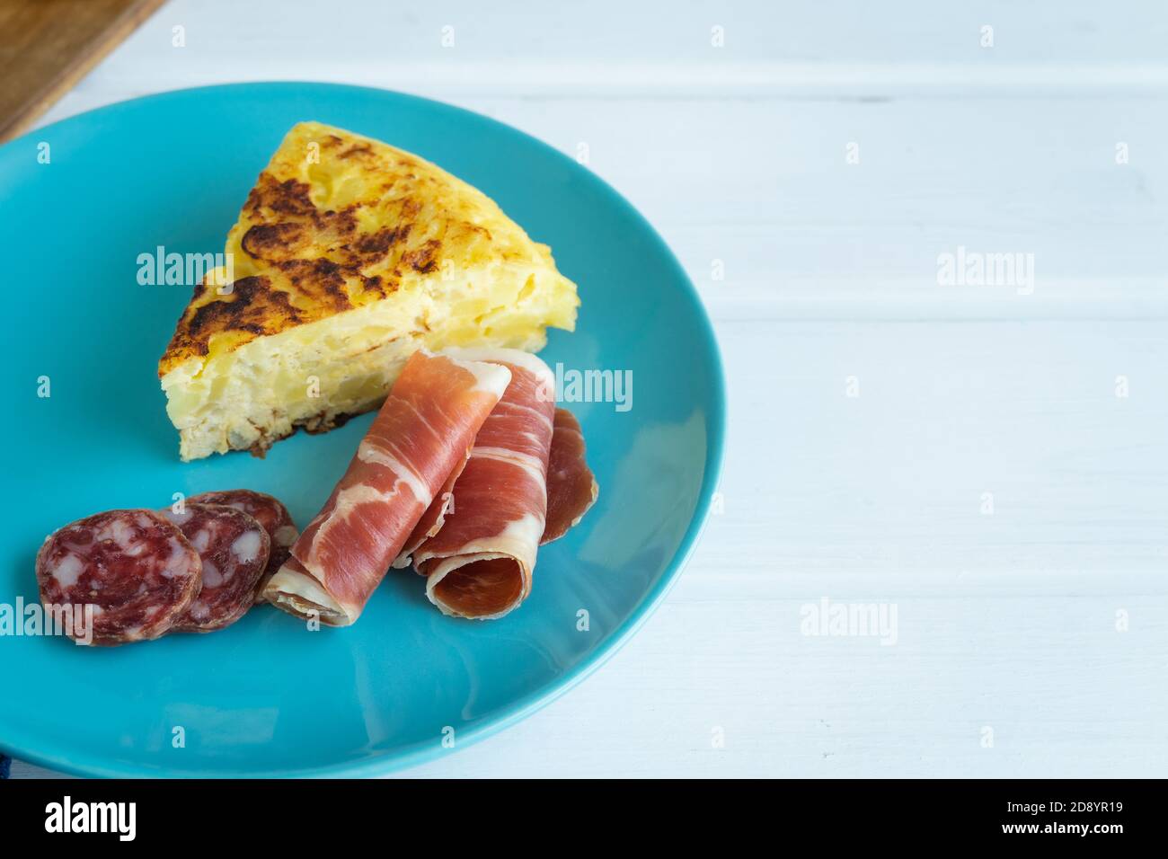 Morceau d'omelette espagnole avec tranches de jambon sur une assiette bleue sur fond blanc. Copiez l'espace vers la droite. Concept alimentaire typique. Banque D'Images