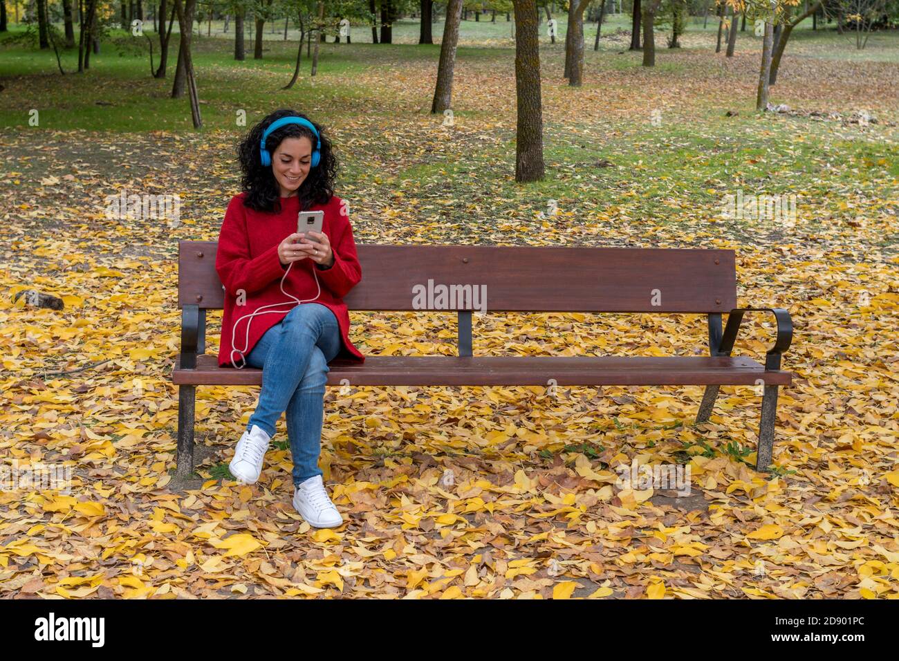 une jeune brunette assise dans un parc à l'écoute de musique avec son téléphone cellulaire et son casque bleu. fond d'arbres colorés et de feuilles mortes Banque D'Images