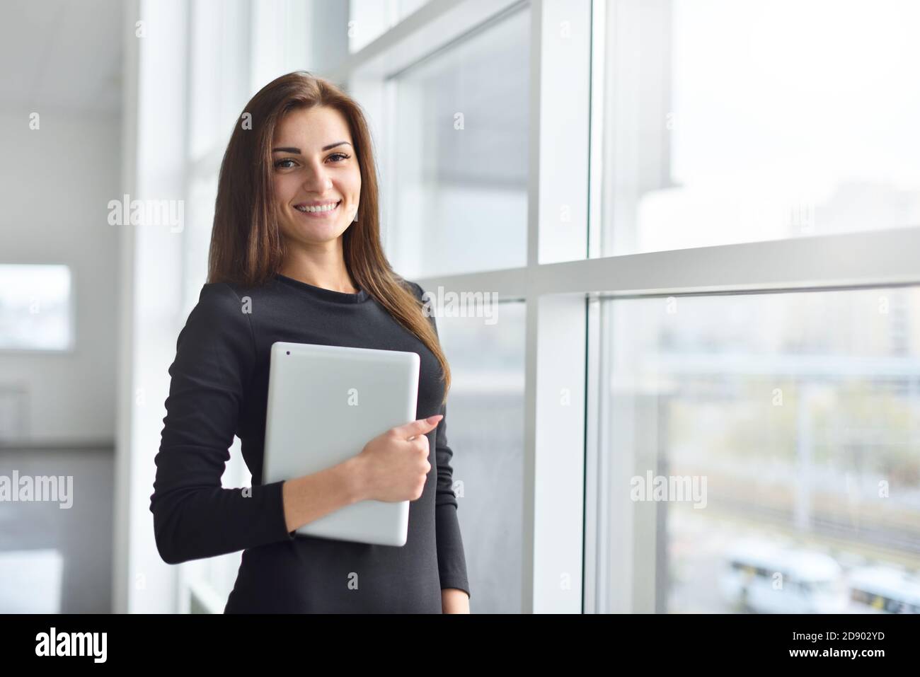 La jeune femme responsable de bureau se tient près de la fenêtre et tient une tablette numérique entre ses mains Banque D'Images